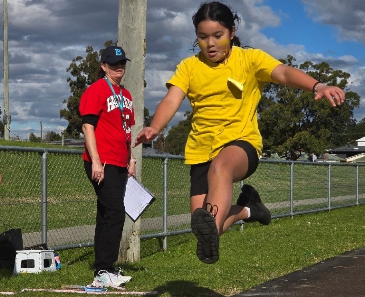 A student jumping high into the air for a long jump event while the teacher stands by to measure the distance
