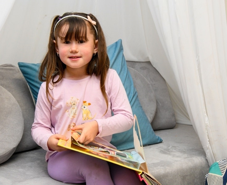 A young girl reading a book on a couch in the preschool.