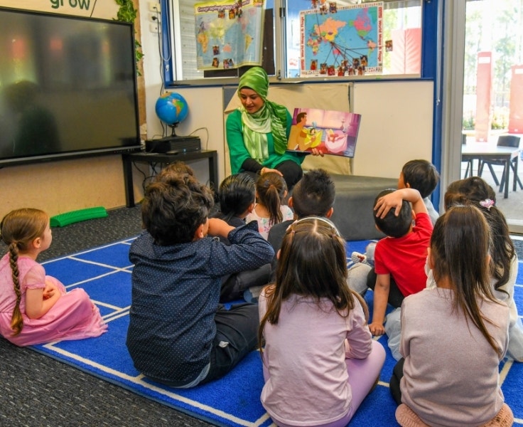 A group of preschool students sitting on the carpet listening to an educator read a picture book.